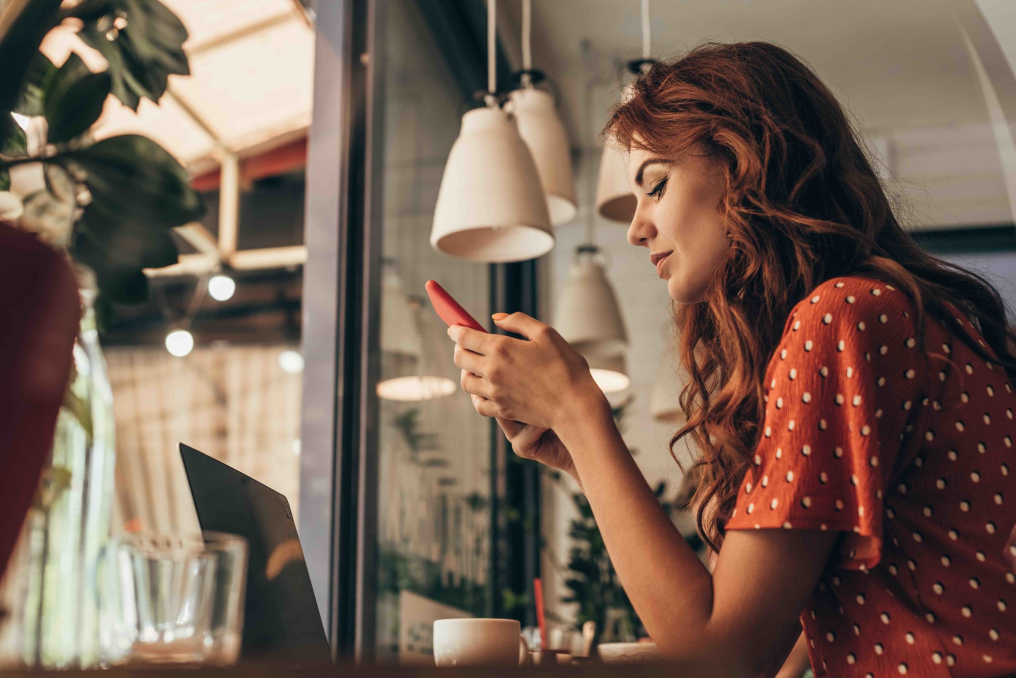 side view of young woman using smartphone at table with laptop in coffee shop