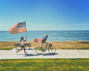 girl chasing a man on bicycle next to shoreline with american flags