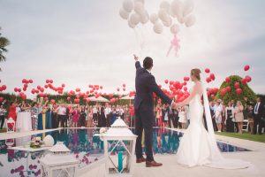 bride and groom standing by a pool, relatives hold red balloons on the other side