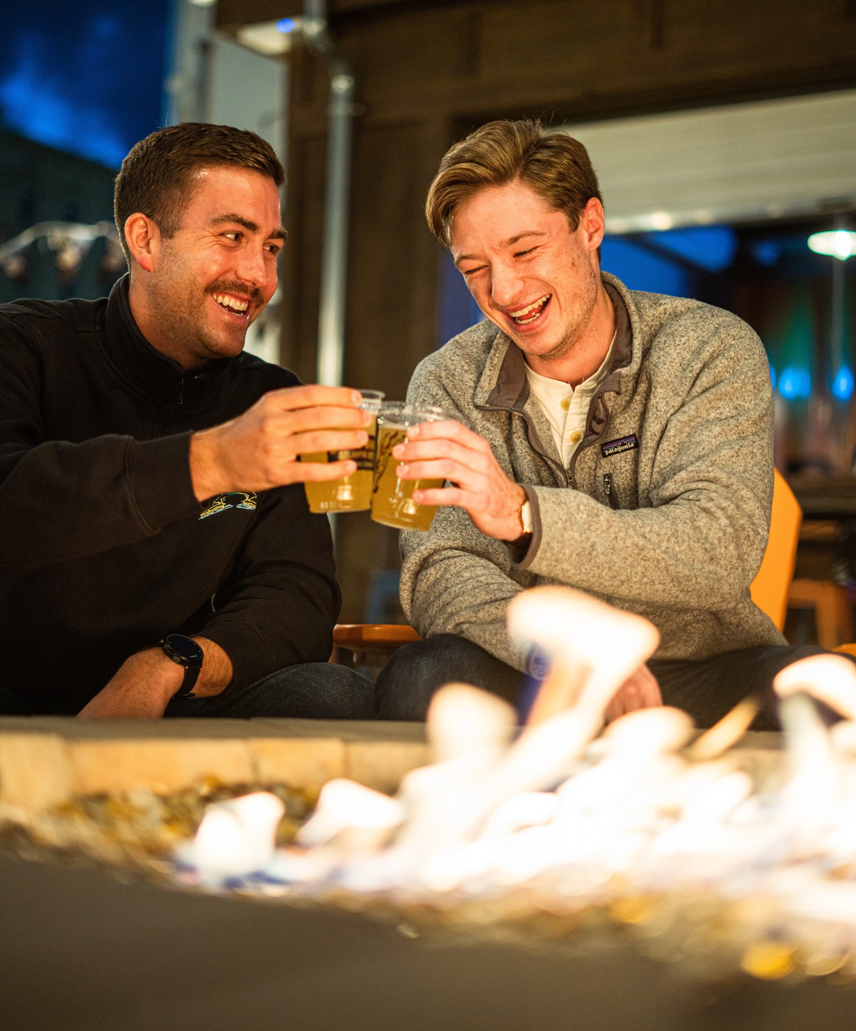 two men drinking beer together by a firepit