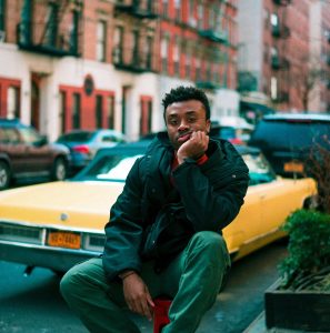 man sitting on trunk of a car on city street looking bored