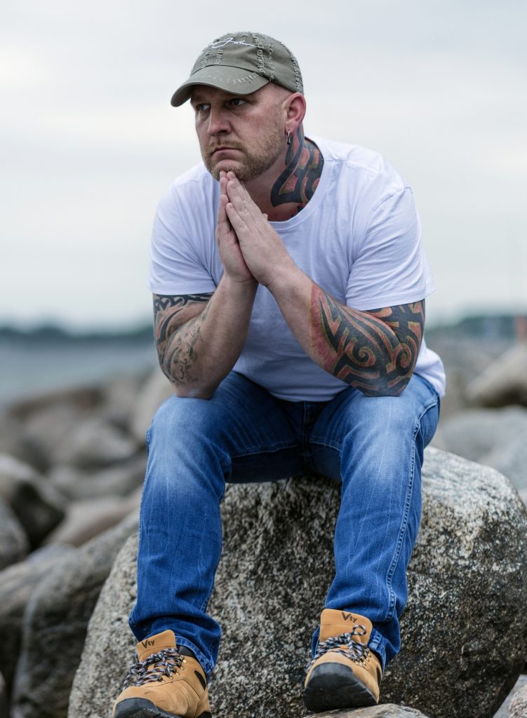tattooed middle aged man sitting on a rock contemplating