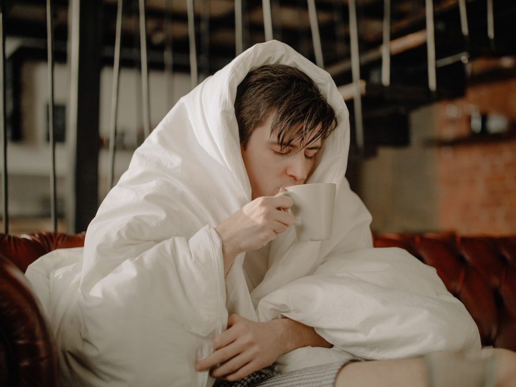 man wrapped in white comforter drinking tea