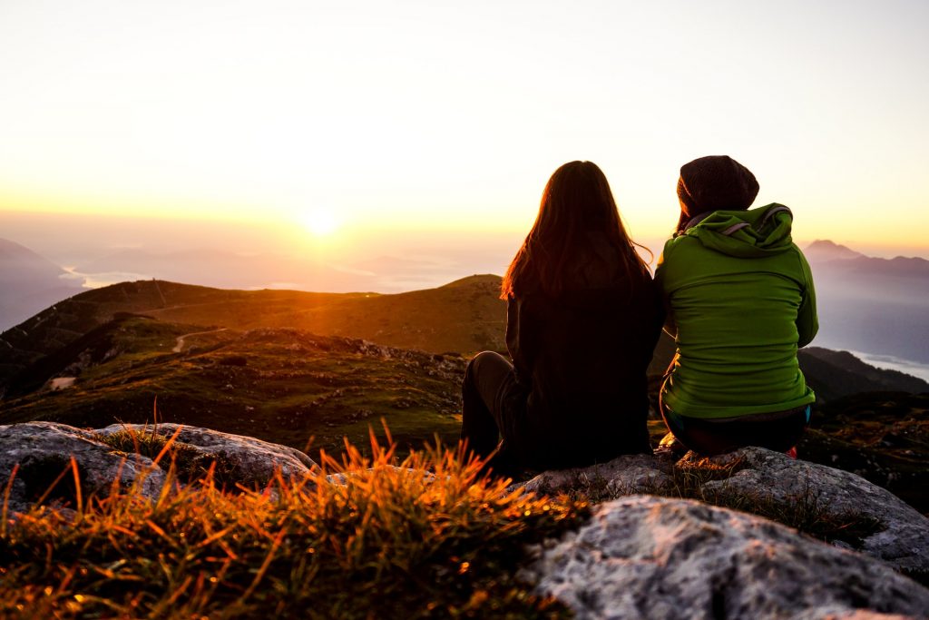 two women sitting on a rock facing the sunset