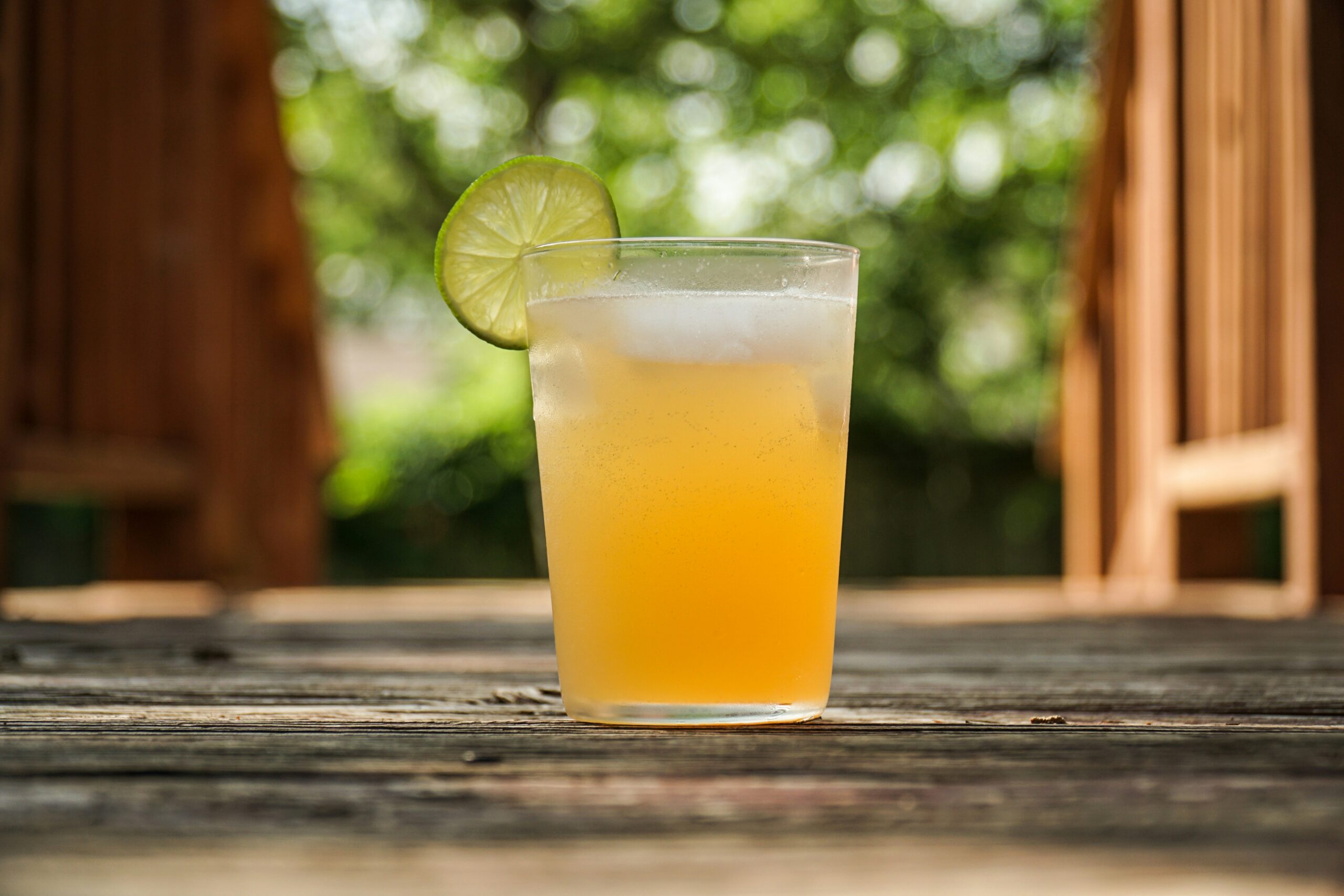 An alcoholic beverage with a lime on the rim of the glass sitting on a wood platform with outdoor greenery in the background.