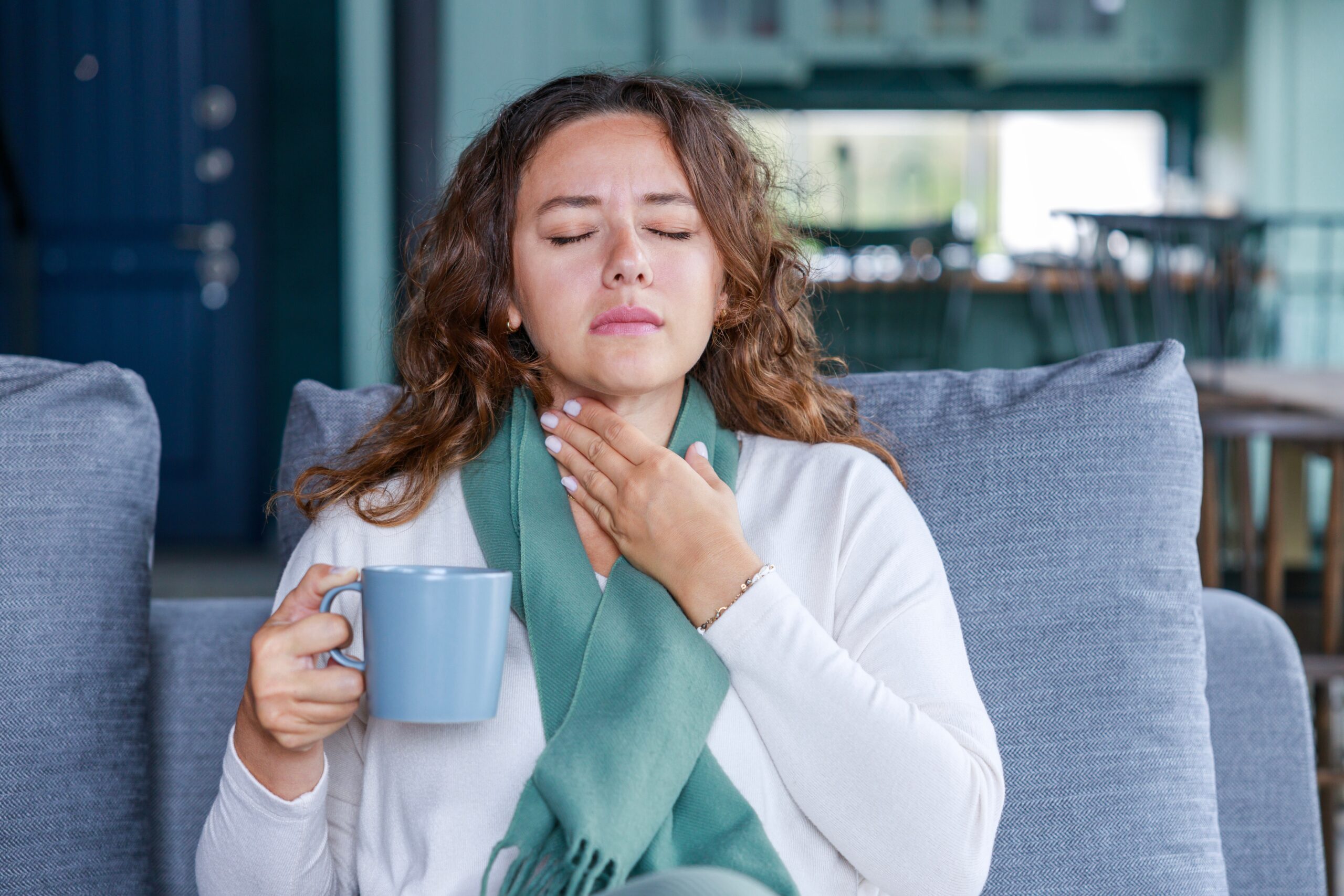 Woman holds a beverage in a blue mug with one hand and holds her throat as though it's sore, with the other hand.